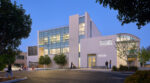 Exterior of the Shiley Eye Institute in La Jolla, California, at dusk, showing a modern multistory medical building with a light facade, large illuminated windows, and a curved roofline on the right. Trees frame the scene, East Entrance signage is visible, and people are near the entrance, including a staff member pushing a patient in a wheelchair.