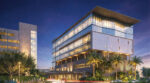 Evening rendering of the Helen and Jacob Shaham Academic Medical Center in Miami, featuring a glass-and-concrete medical building with a covered entrance, illuminated interiors, tropical landscaping, and an adjacent parking garage.