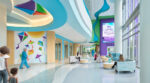 The main lobby of Mary Bridge Children's Hospital featuring a 3D kite ceiling sculpture, kite-themed murals, wave-patterned terrazzo floors, blue armchairs and a commemorative digital display, where families and staff move through the space.