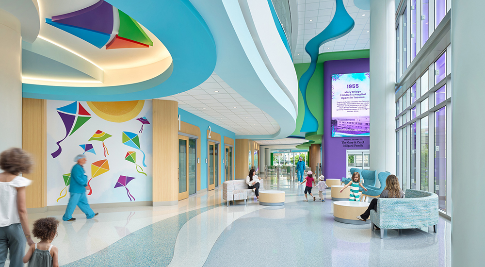 The main lobby of Mary Bridge Children's Hospital featuring a 3D kite ceiling sculpture, kite-themed murals, wave-patterned terrazzo floors, blue armchairs and a commemorative digital display, where families and staff move through the space.